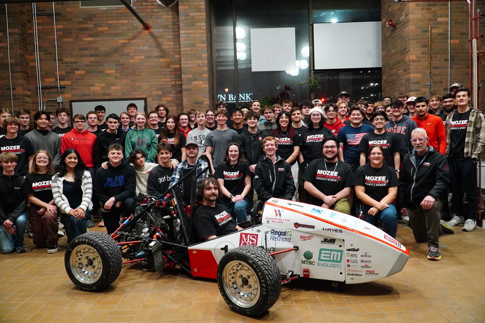 The full Mozee Motorsports team, more than 50 students, posing with the number 1 hybrid formula car inside an MSOE building